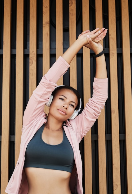 African young woman listening to music in wireless headphones raising her arms and exercising during...