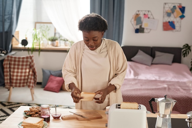 Giovane donna africana che fa i toast con marmellata per colazione in cucina