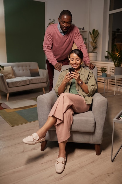 African young woman sitting on armchair looking photos on her mobile phone with man standing behind her in the room