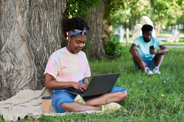 African young woman sitting on the grass and working online on laptop in the park