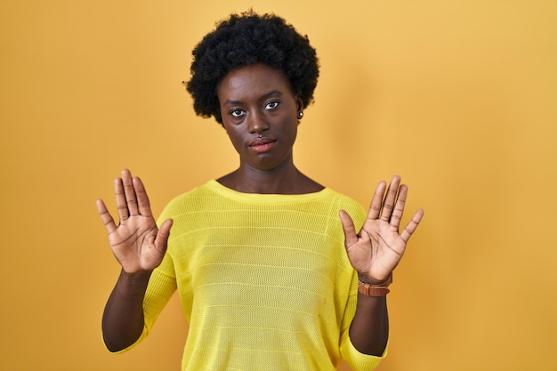 Photo african young woman standing over yellow studio moving away hands palms showing refusal and denial with afraid and disgusting expression stop and forbidden