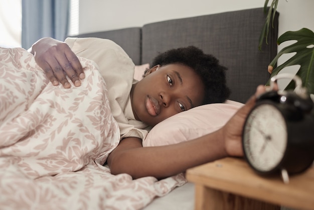 ベッドに横になって目覚まし時計を消しているアフリカの若い女性、彼女は朝目を覚ます