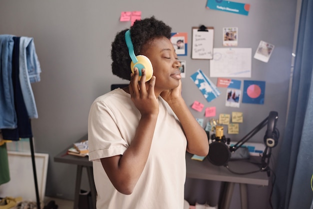 African young woman with closed eyes enjoying the music in wireless headphones while standing in the room