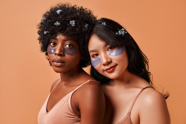 Africanamerican and asian young women using eye patches in lingerie posing on beige background cosmetology and spa treatments for face and body