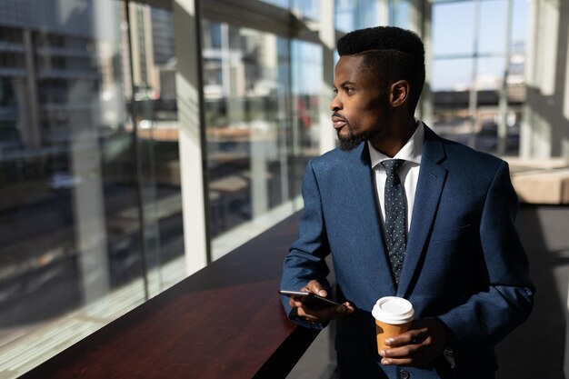 Foto uomo d'affari afroamericano con una tazza di caffè e un telefono cellulare che guarda lontano in ufficio