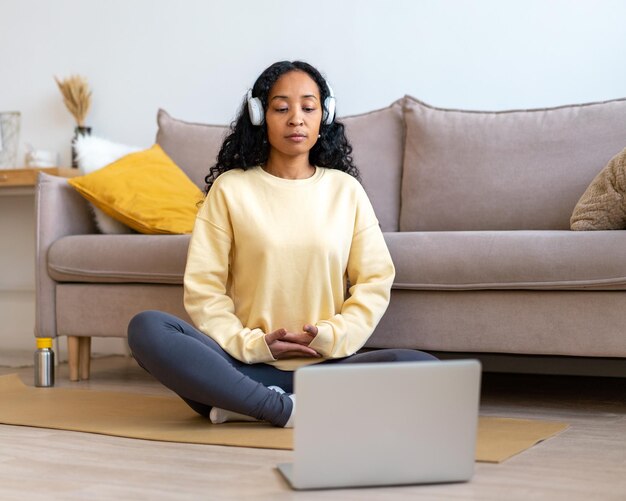 Africanamerican female listening to music in headphones while meditating with laptop at home