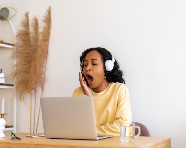 Africanamerican female student yawning while listening to online lecture in headphones on laptop