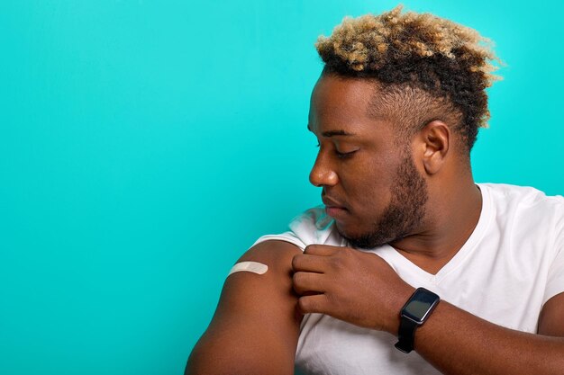 AfricanAmerican guy after vaccination looks at his shoulder with a patch on a turquoise background