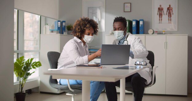 AfricanAmerican male doctor and patient in protective masks talking at clinic using laptop