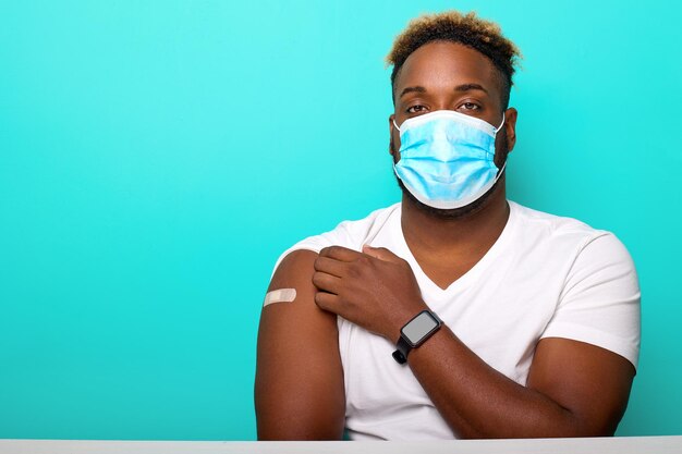 AfricanAmerican man in a mask sitting at a table shows his shoulder with a patch after vaccination having received a coronavirus vaccine on a turquoise background Concept of vaccination