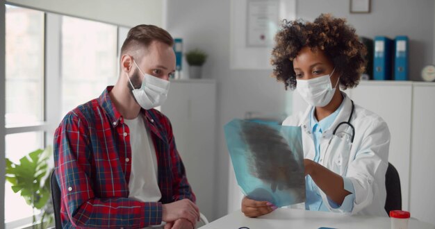 Foto medico donna afroamericana in maschera di sicurezza che mostra i raggi x al paziente presso l'ufficio medico in ospedale