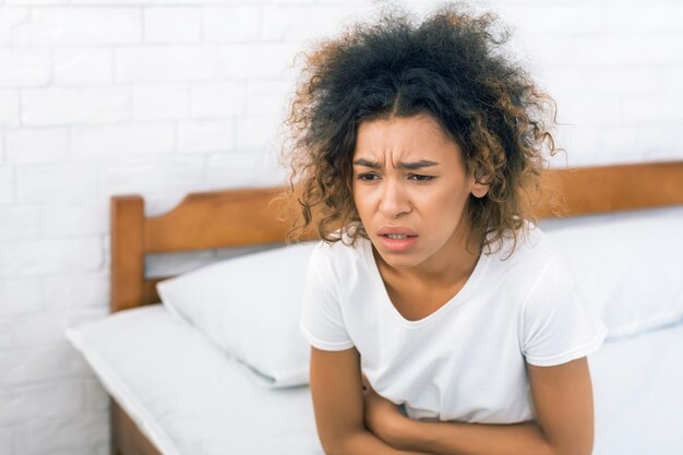 Photo africanamerican woman having painful stomachache sitting on bed