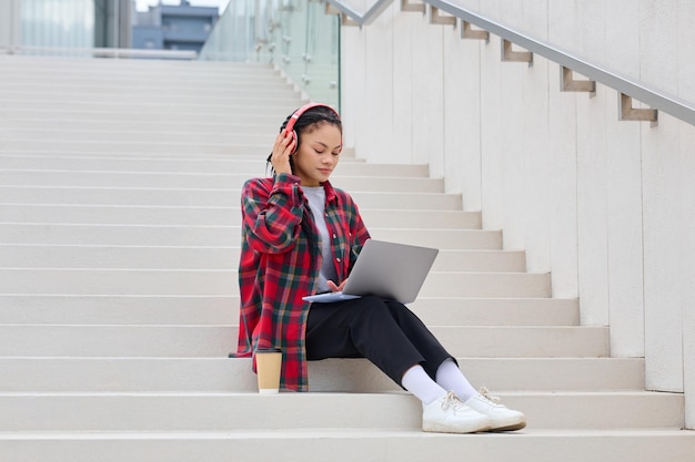 An AfricanAmerican woman is sitting on the steps and working on a laptop listening to music with headphones Freelancing and working outside the office