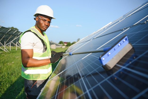 AfricanAmerican worker in gray overalls and a white hard hat works in a field of solar panels Solar renewable energy concept