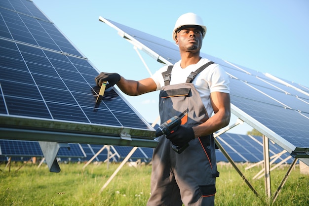 灰色のオーバーオールと白いハードヘットを着たアフリカ系アメリカ人の労働者が太陽電池パネルの畑で働いています 太陽の再生可能エネルギーコンセプト