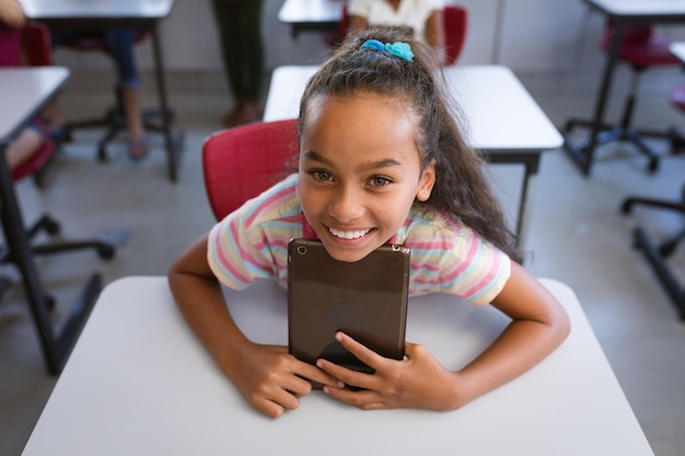 Afrikaans-Amerikaans meisje dat glimlacht terwijl ze een digitale tablet op haar bureau houdt in de klas op school