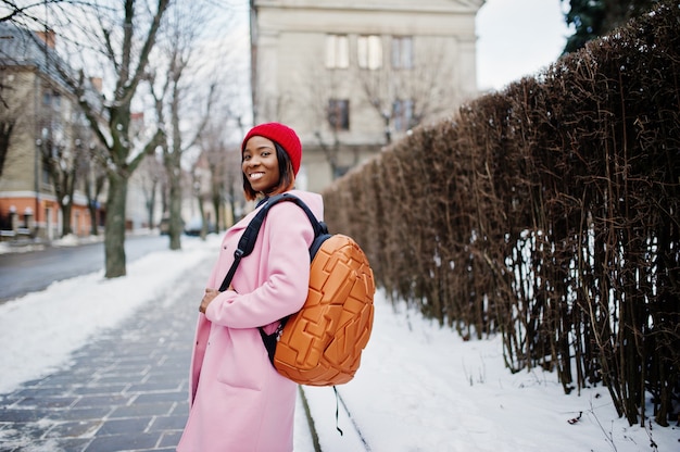 Afrikaans amerikaans meisje in rode hoed en roze laag met rugzak bij straat van stad op de winterdag.