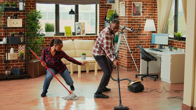 Afrikaans-Amerikaans paar dansen en appartementkamers schoonmaken, dweil gebruiken om vuil te wassen en stofzuigen om vloeren schoon te maken. Jonge levenspartners genieten van de voorjaarsschoonmaak met wasoplossing.