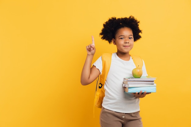 Afrikaans-Amerikaans schoolmeisje dat notitieboekjes en boeken vasthoudt en iets laat zien op gele achtergrondkopieerruimte