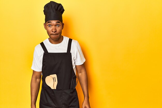 Afrikaans-Amerikaanse kok met gereedschap gele studio verward voelt zich twijfelachtig en onzeker