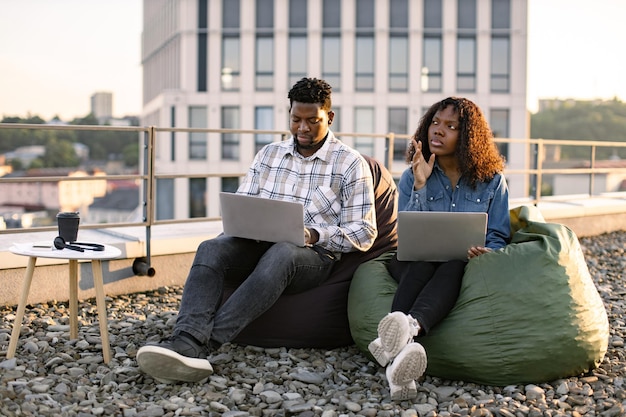 Afrikaans echtpaar met laptops terwijl ze in bonenzakken op het dak van een kantoor zitten