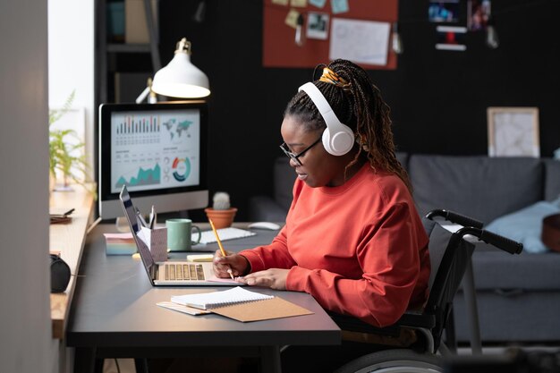 Afrikaans meisje met een draadloze koptelefoon zit aan de tafel voor de laptop en maakt aantekeningen, ze studeert thuis