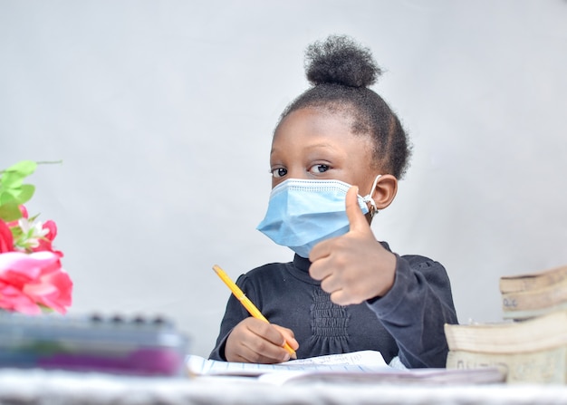 Afrikaans meisjeskind met neusmasker dat duimen opgeeft tijdens het studeren en lezen van haar boeken