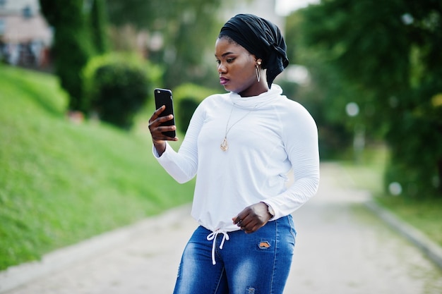 Afrikaans moslimmeisje in zwarte hijab witte sweater en jeans poseerde buiten met mobiele telefoon