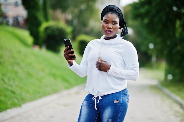 Afrikaans moslimmeisje in zwarte hijab witte sweater en jeans poseerde buiten met mobiele telefoon
