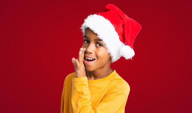 Afrikaanse Amerikaanse jongen die met Kerstmishoed iets over rode achtergrond fluistert