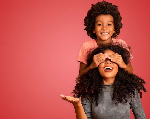 Afrikaanse Amerikaanse jongen die zijn moeder gesloten ogen houdt. Rode achtergrond. Moederdag. Braziliaanse familie.