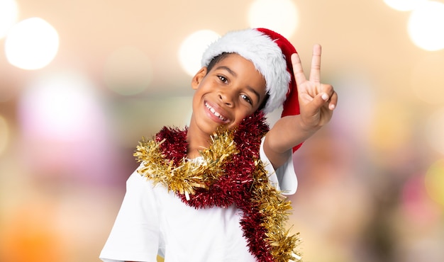 Afrikaanse amerikaanse jongen met kerstmishoed die en overwinningsteken over vage muur glimlachen tonen
