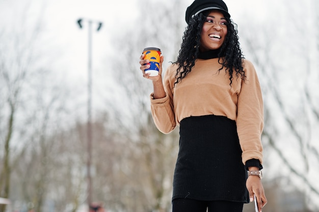 Afrikaanse amerikaanse vrouw in zwarte rok, bruine sweater en glb gesteld bij de winterdag tegen sneeuwachtergrond, die kop van koffie houden