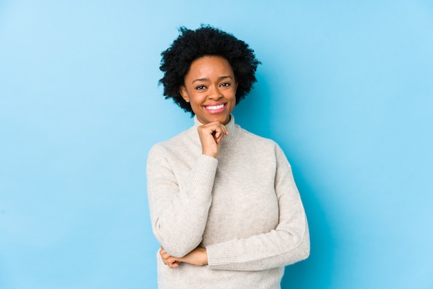 Afrikaanse Amerikaanse vrouw van middelbare leeftijd tegen een blauwe muur die gelukkig en zeker, wat betreft kin met hand glimlacht.