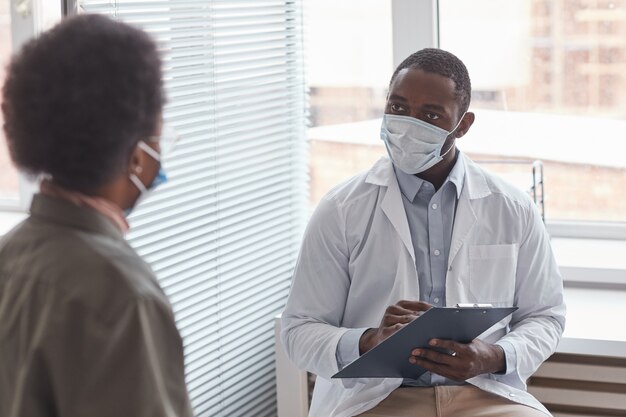 Afrikaanse arts met beschermend masker in gesprek met zijn patiënt tijdens haar bezoek aan het ziekenhuis