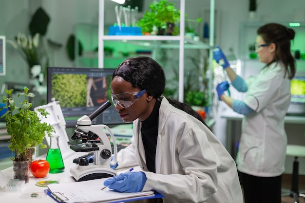 Afrikaanse biochemie arts die chemische test onderzoekt met behulp van een microscoop voor genetische onderzoeker. Bioloog-specialist ontdekt biologische ggo-planten tijdens het werken in een microbiologisch voedsellaboratorium.