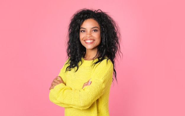 Afrikaanse etnische vrouw met gitzwart golvend haar in geel gebreid sweatshirt poseert terwijl ze naar de camera kijkt en lacht van vreugde