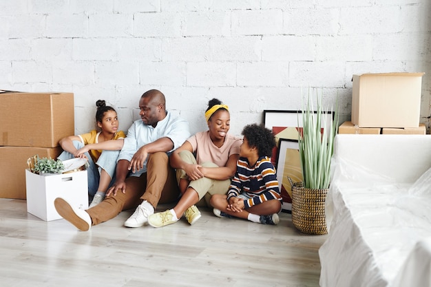 Afrikaanse familie van vader, moeder, zoontje en tienerdochter die plannen bespreken terwijl ze thuis op de vloer tegen een wit geschilderde muur zitten