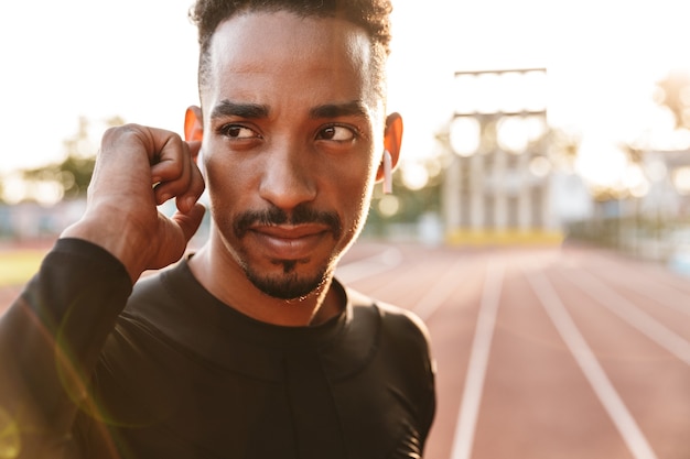 afrikaanse geconcentreerde serieuze knappe jonge sportman in het stadion die buiten muziek luistert met draadloze bluetooth-oortelefoons.