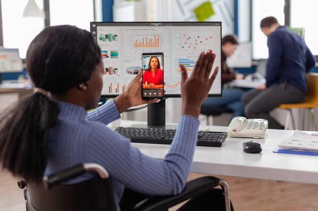 Afrikaanse gehandicapte werknemer zwaait naar online partner tijdens videogesprek zittend geïmmobiliseerd in rolstoel...