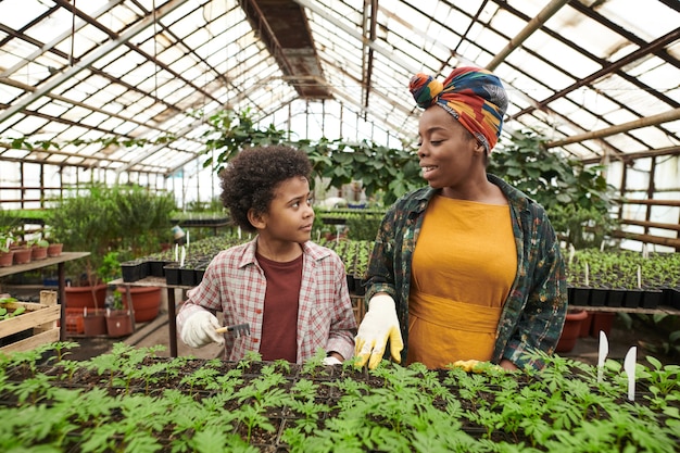 Afrikaanse gelukkige vrouw die met haar kind praat terwijl ze planten in de kas planten