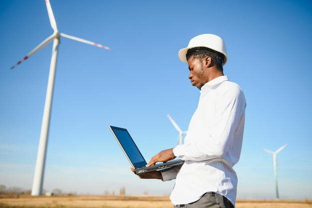 Afrikaanse ingenieur die witte bouwvakker draagt die zich met digitale tablet tegen windturbine bevindt