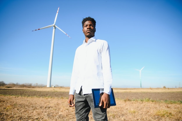 Afrikaanse ingenieur met witte helm die op zonnige dag met digitale tablet tegen windturbine staat