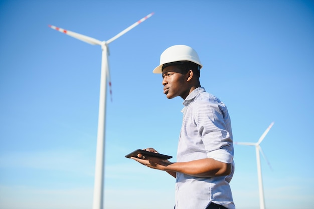 Afrikaanse ingenieur met witte helm die op zonnige dag met digitale tablet tegen windturbine staat