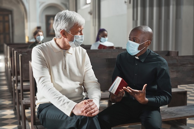 Afrikaanse jonge priester met masker in gesprek met senior man terwijl ze op de bank in de kerk zitten