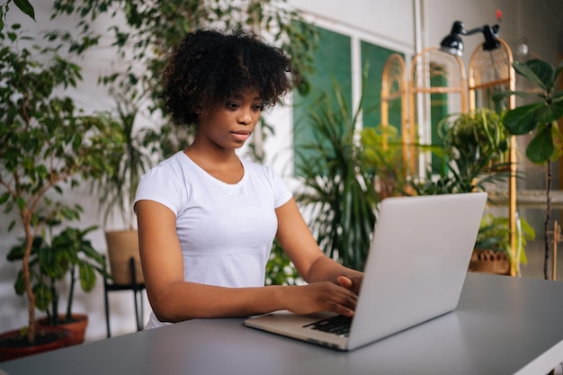 Afrikaanse jonge vrouw die aan het typen is op een laptopcomputer die aan een bureau zit in een kantoor aan huis met een modern biofilie-ontwerp