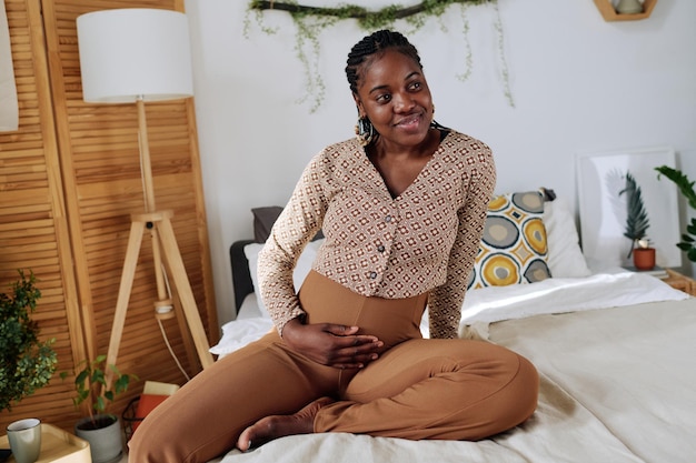 Afrikaanse jonge vrouw die haar zwangere buik aanraakt en glimlacht terwijl ze in haar slaapkamer zit