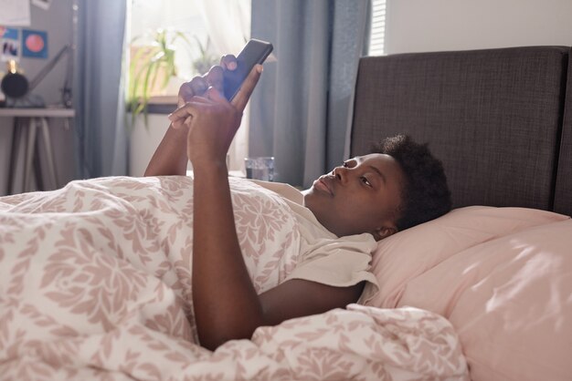 Afrikaanse jonge vrouw die onder de deken op het bed ligt en online leest op mobiele telefoon in de slaapkamer