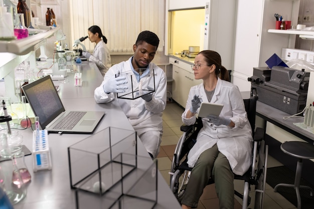 Afrikaanse jonge wetenschapper die muis in de doos houdt met zijn collega die in een rolstoel zit en digitale tablet gebruikt in het laboratorium