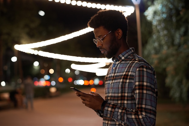 Afrikaanse jongeman leest een bericht online op mobiele telefoon terwijl hij 's avonds in de stad staat